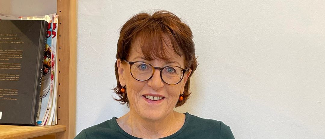 Eine ältere Frau mit kurzen Haaren und einer Brille sitzt auf einem Stuhl und hält ein offenes Buch in der Hand. Sie lächelt und scheint für ein Foto zu posieren. Hinter ihr steht ein Holzregal mit mehreren Büchern darauf.