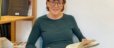 Eine Frau mit kurzen Haaren und Brille sitzt in einem Stuhl, hält ein offenes Buch und lächelt. Hinter ihr steht ein Holzregal mit mehreren Büchern. Die Wand hinter ihr ist weiß.