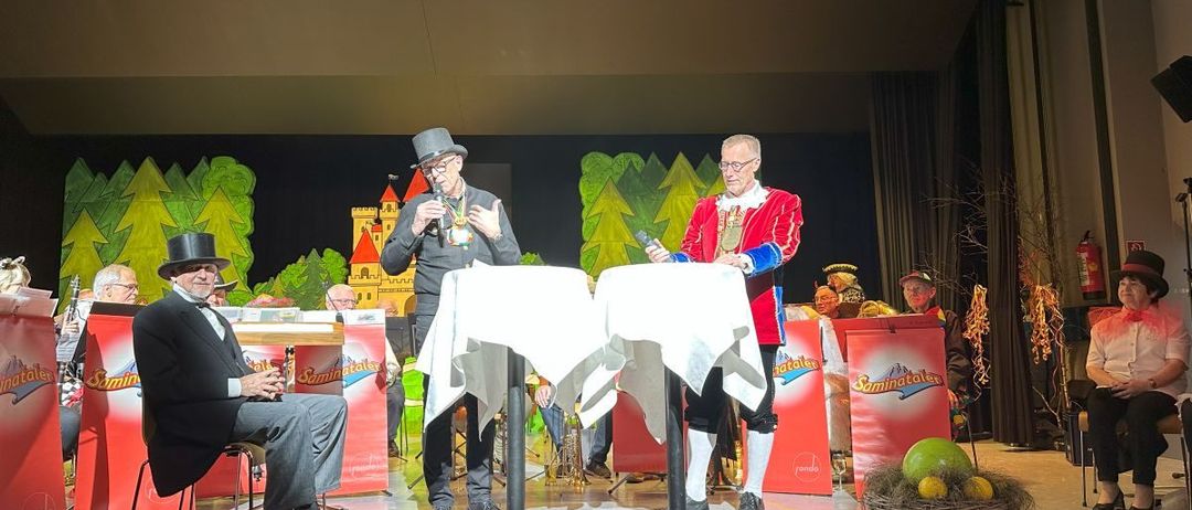 Drei Männer auf einer Bühne in Kostümen. Einer hält ein Mikrofon und ein anderer raucht eine Zigarre. Ein Korb mit Obst steht auf der Bühne.