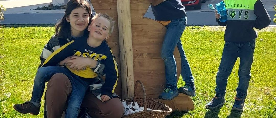 Drei Kinder posieren neben einem riesigen Holzhasen; einer steht auf dem Hasen, ein anderer hält einen Korb und der dritte Junge hält eine Wasserflasche. Die Szene ist im Freien mit Gebäuden und Autos im Hintergrund.