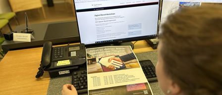 Eine Person sitzt vor einem Schreibtisch und konzentriert sich auf einen Computerbildschirm, der 'Digital Ueberall Workshops' anzeigt. Der Bildschirm zeigt eine Webseite mit Informationen und ein Flyer in der Nähe.
