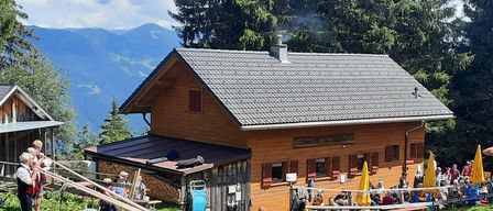 Ein Holzchalet mit grauem Dach und Schornstein, umgeben von einer Gruppe von Menschen, die auf einer Bank unter einem gelben Regenschirm sitzen.