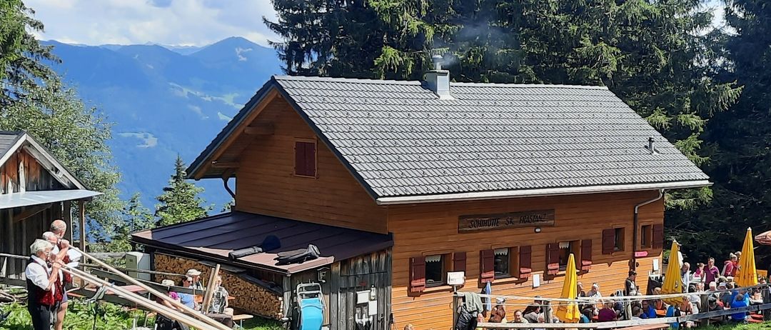 Ein Holzchalet mit grauem Dach und Schornstein, umgeben von einer Gruppe von Menschen, die auf einer Bank unter einem gelben Regenschirm sitzen.