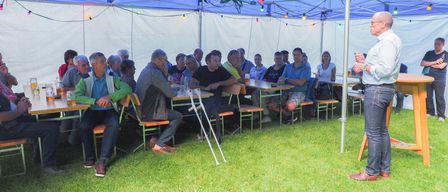 Eine Gruppe von Menschen sitzt unter einem blauen Zelt mit Tischen und Stühlen, einige hören einem Mann zu, der spricht. Das Zelt ist mit Lichterketten dekoriert.