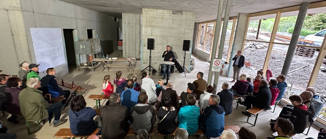 Eine Gruppe von Menschen ist in einem Halbkreis um einen Mann versammelt, der an einem Podium mit Mikrofon steht. Der Raum hat Betonwände und große Fenster. Einige Leute sitzen auf Bänken, während andere stehen. Es gibt zwei Lautsprecher auf Ständern und ein Schild in der Nähe der Fenster.