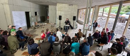 Eine Gruppe von Menschen ist in einem Halbkreis um einen Mann versammelt, der an einem Podium mit Mikrofon steht. Der Raum hat Betonwände und große Fenster. Einige Leute sitzen auf Bänken, während andere stehen. Es gibt zwei Lautsprecher auf Ständern und ein Schild in der Nähe der Fenster.