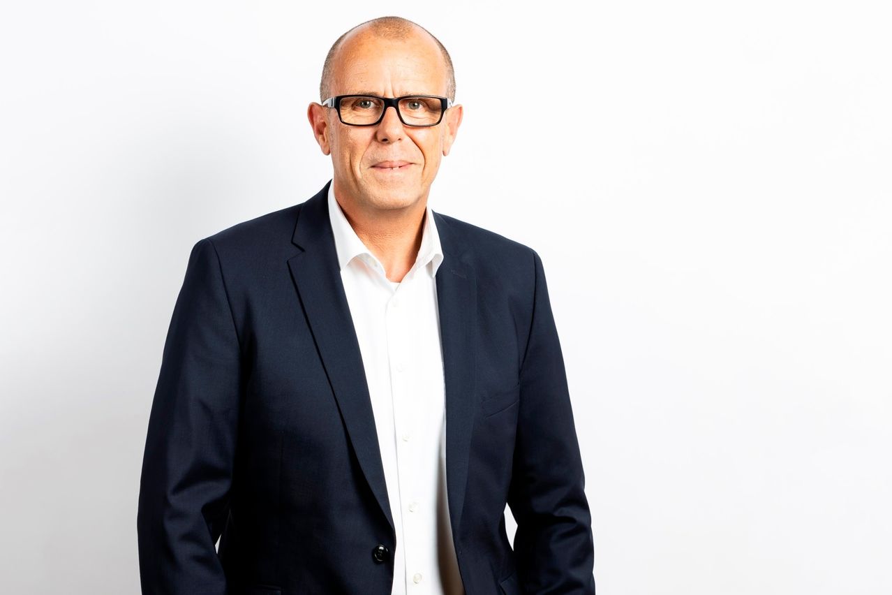 Ein Mann in einem dunkelblauen Blazer über einem weißen Hemd steht vor einem weißen Hintergrund. Er lächelt, trägt eine Brille und hat einen leichten Schatten auf seiner rechten Seite.