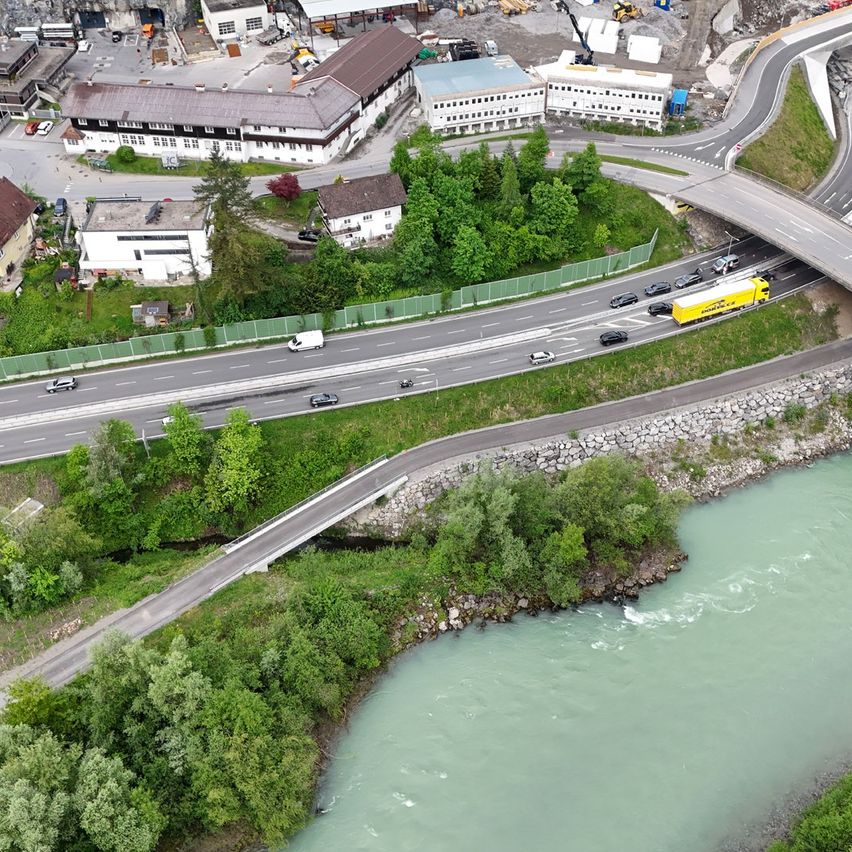 Eine Luftaufnahme einer Autobahn mit Fahrzeugen, die vorbeifahren, in der Nähe eines Flusses. Gebäude und Baustellen sind im Hintergrund zu sehen.