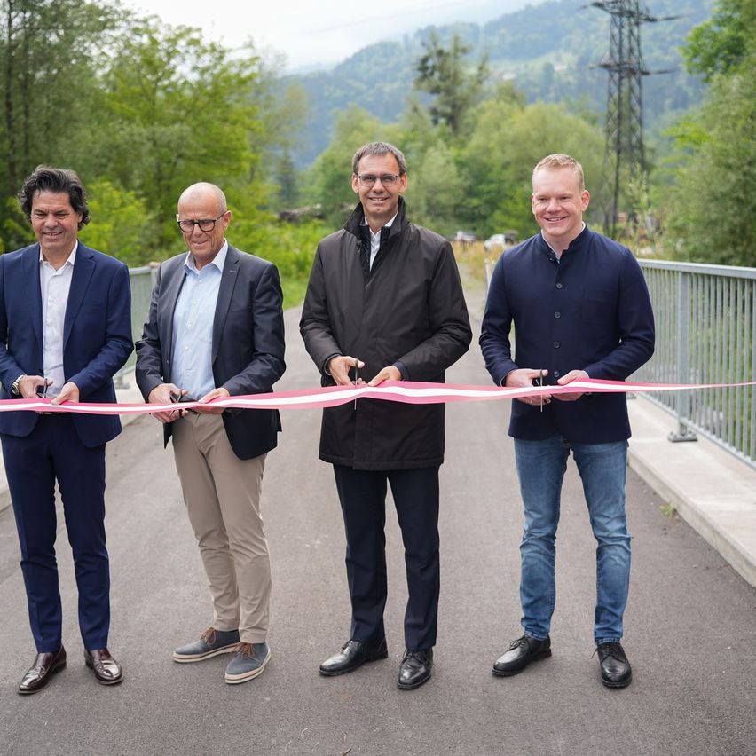 Vier Männer in Anzügen stehen auf einer Brücke und halten ein Band, möglicherweise für eine Schleifenschneidezeremonie. Hinter ihnen ist ein Metallgeländer, und im Hintergrund sind Bäume und Berge.