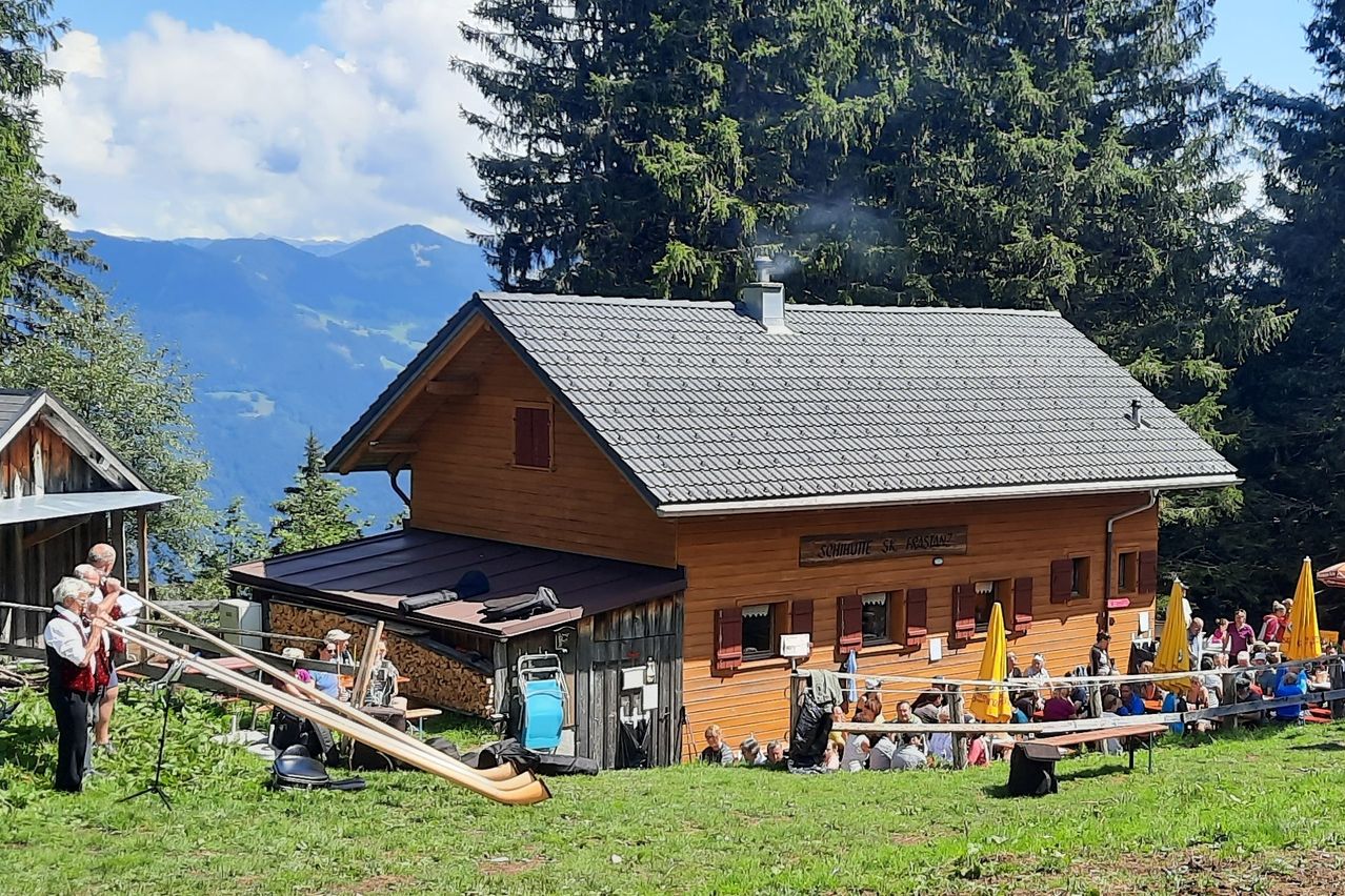Ein Holzchalet mit Schornstein steht vor einem Wald. Es sitzen viele Menschen um einen Tisch, und einige stehen. Es gibt Tische und Stühle draußen.