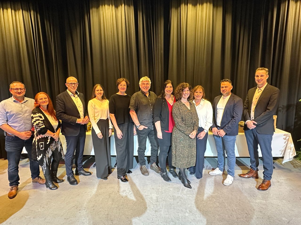 Eine Gruppe von Menschen steht vor einem schwarzen Vorhang und lächelt für ein Foto. Sie sind in formeller Kleidung gekleidet.