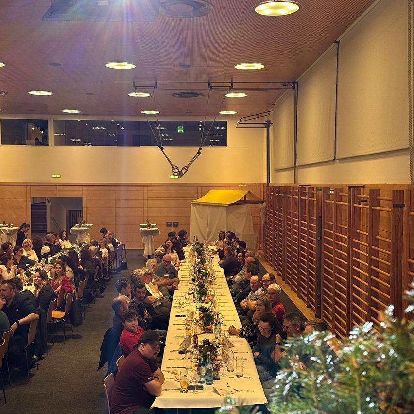 Eine große Indoor-Veranstaltung zeigt einen langen Tisch mit vielen sitzenden Gästen, geschmückt mit Pflanzen und Gläsern. Der Raum hat Holzwände, ein gelbes Zelt und große Fenster.