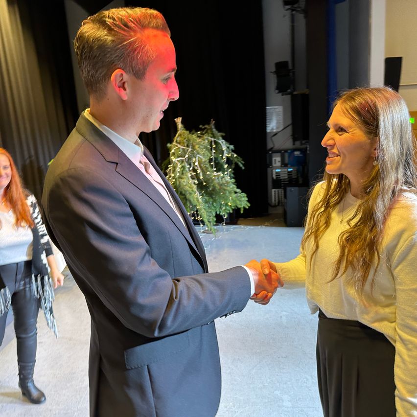 Ein Mann in einem Anzug und eine Frau in einem Pullover schütteln sich die Hände in einem Raum mit einer Pflanze und einem Lautsprecher. Eine Frau in einem Pullover und Stiefeln steht im Hintergrund.