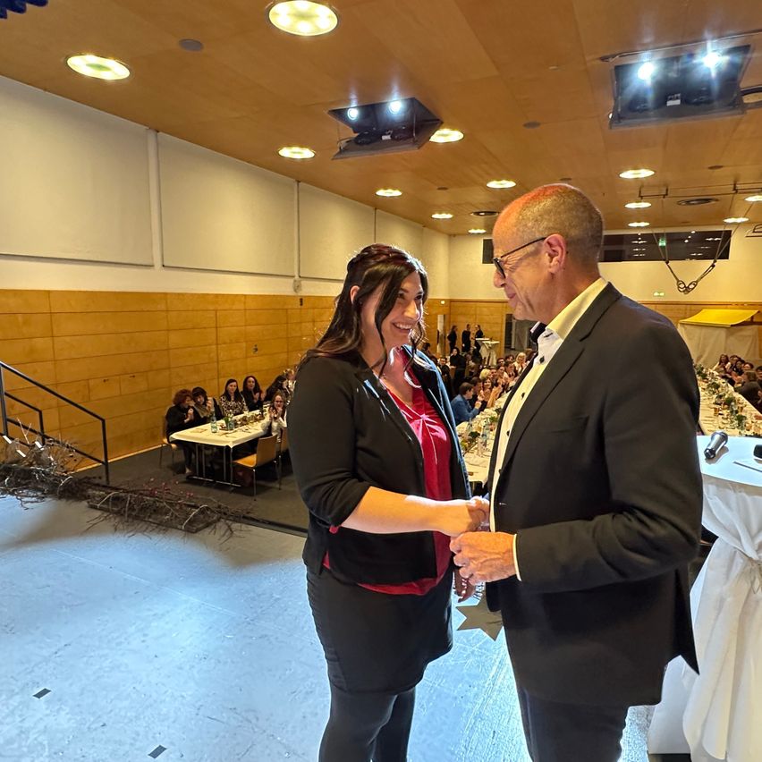 Eine Frau und ein Mann schütteln sich in einem Raum bei einer Veranstaltung die Hände. Die Frau lächelt und der Mann trägt eine Brille. Im Hintergrund befinden sich Tische und Stühle.