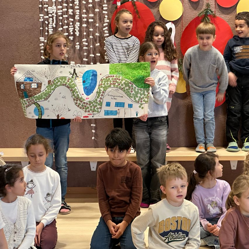 Eine Gruppe von Kindern steht auf einer Bühne und hält ein Plakat. Das Plakat hat eine Zeichnung einer Landschaft. Die Kinder tragen Winterkleidung und einige tragen eine Brille. Hinter ihnen ist eine Wand mit roten und gelben Kreisen verziert.