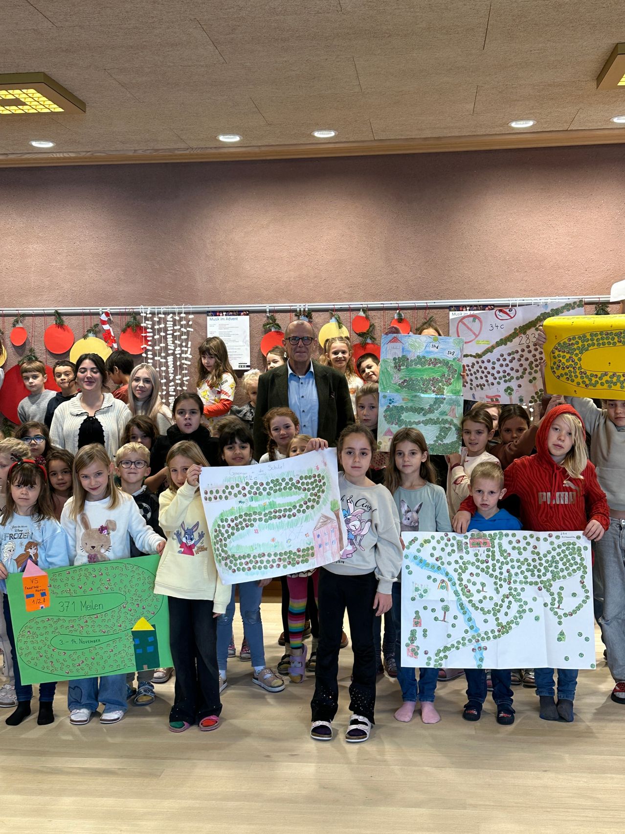 Eine Gruppe von Kindern und Erwachsenen steht in einem Raum und hält Zeichnungen hoch und lächelt. Ein Mann im Anzug steht in der Mitte. Papiere und Dekorationen hängen an der Wand hinter ihnen.