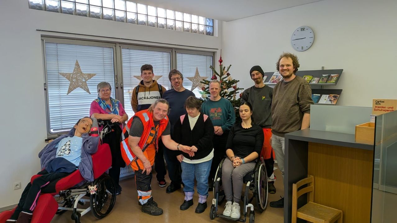 Eine Gruppe von Menschen in einem Raum mit Weihnachtsdekoration. Einige sitzen in Rollstühlen, andere stehen dahinter. Ein Mann in einer orangefarbenen Weste schüttelt einer Frau im Rollstuhl die Hand.