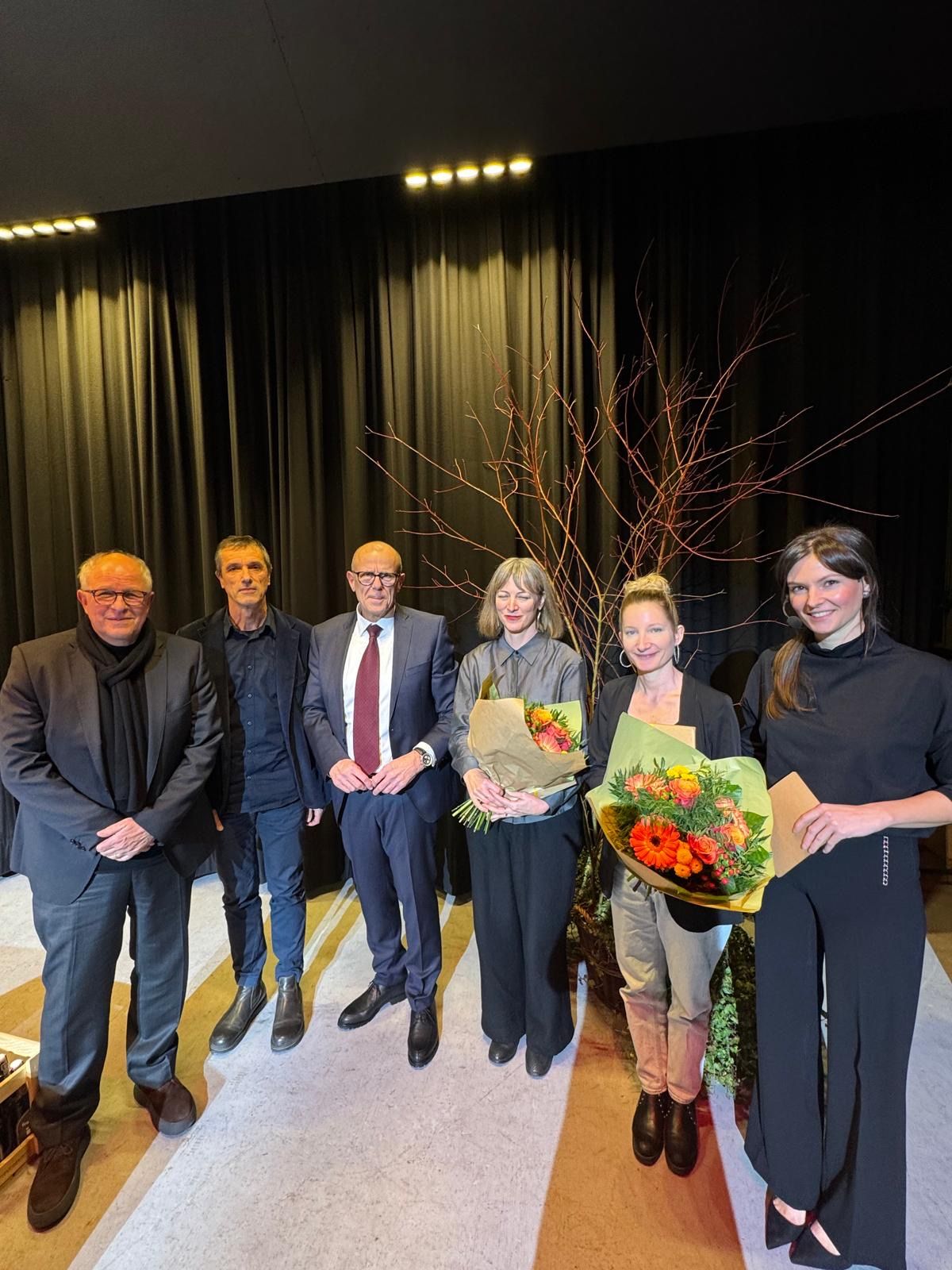 Fünf Personen stehen zusammen vor einer Bühne mit schwarzem Vorhang im Hintergrund. Sie sind alle in formeller Kleidung gekleidet. Eine Frau hält einen Blumenstrauß.