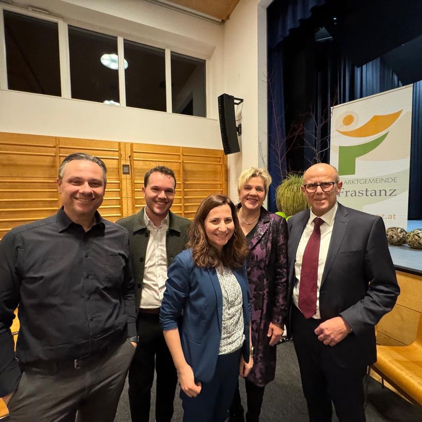 Fünf Personen posieren für ein Foto in einem Raum. Drei Männer und zwei Frauen, alle lächelnd. Die Frau in der Mitte trägt einen blauen Blazer. Hinter ihnen steht ein Banner mit der Aufschrift 'Marktgemeinde Frastanz.' Holzpaneele säumen die Wand, und ein Lautsprecher ist montiert.