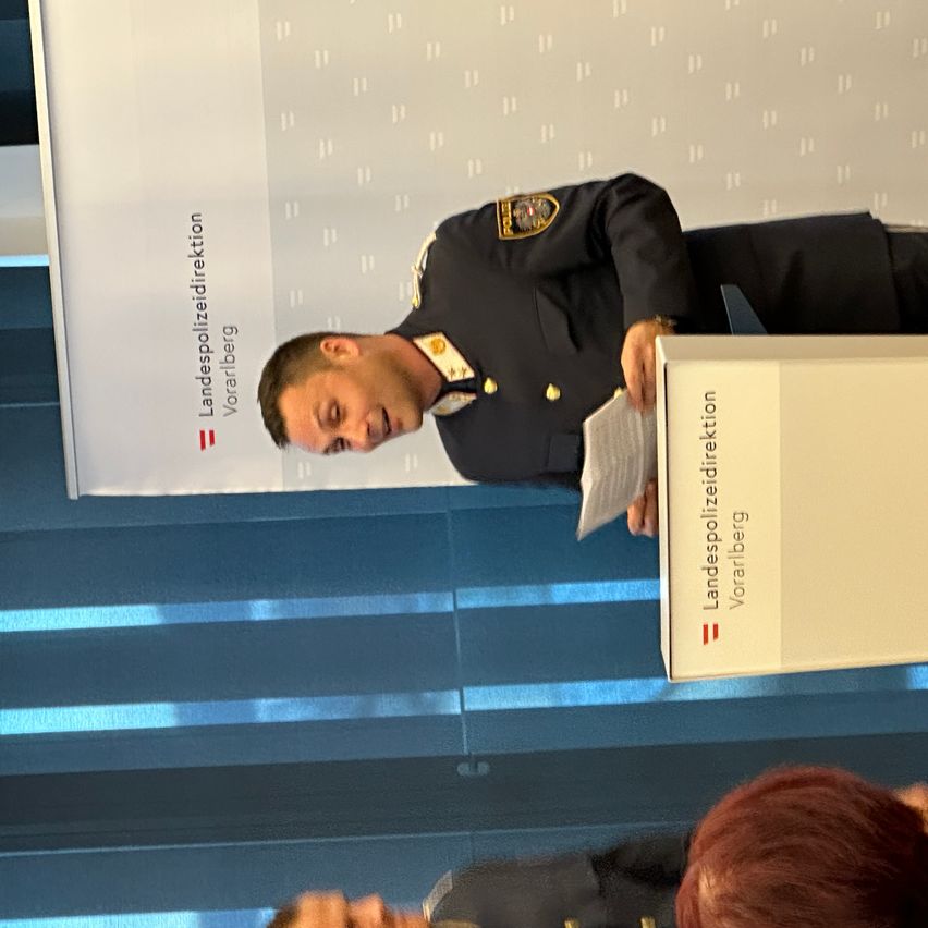 Ein Polizist steht an einem Podium mit einer Box, die 'Landespolizeiinspektion Vorarlberg' beschriftet ist. Er hält ein Papier und scheint an ein Publikum zu sprechen.