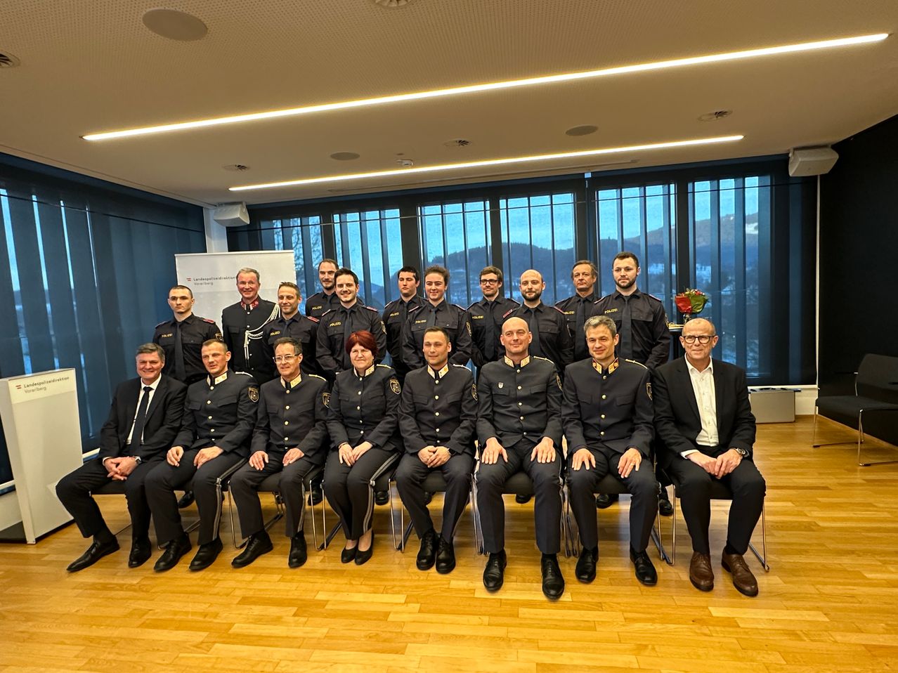 Eine Gruppe uniformierter Personen, wahrscheinlich Polizeibeamte, posiert für ein Foto in einem Raum mit Holzböden und großen Fenstern.