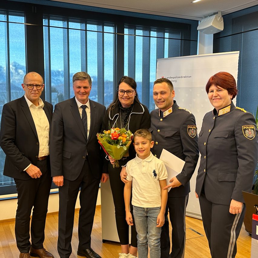 Fünf Erwachsene und ein junger Junge stehen vor einer Wand. Der Junge hält einen Blumenstrauß. Die Erwachsenen sind in Anzügen und Uniformen gekleidet.