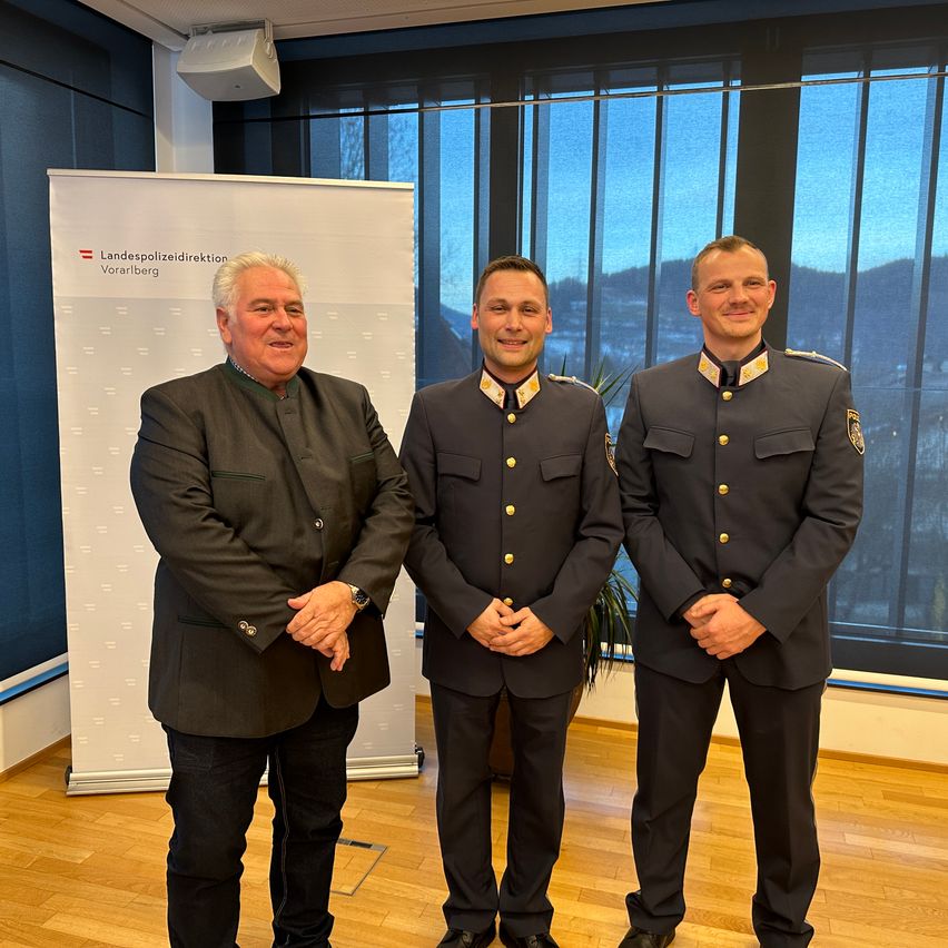 Drei Männer stehen nebeneinander vor einem großen Fenster. Ein Mann ist in einem formellen Anzug, und die anderen beiden sind in Militäruniformen. Hinter ihnen steht ein Banner, das 'Landespolizeidirektion Vorarlberg' besagt.