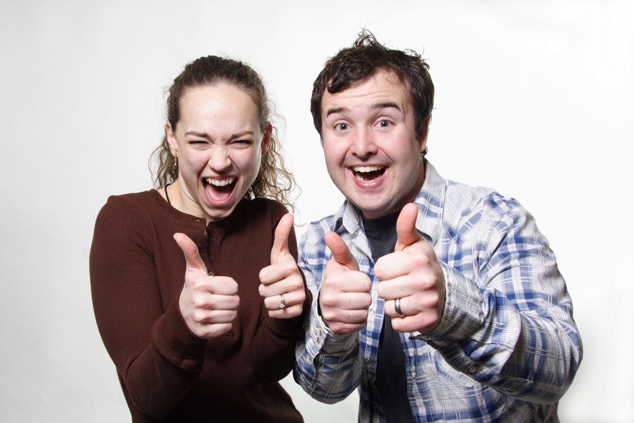 Eine Frau und ein Mann stehen eng beieinander und zeigen beide Daumen hoch. Die Frau lächelt mit offenem Mund, und der Mann lächelt breit.