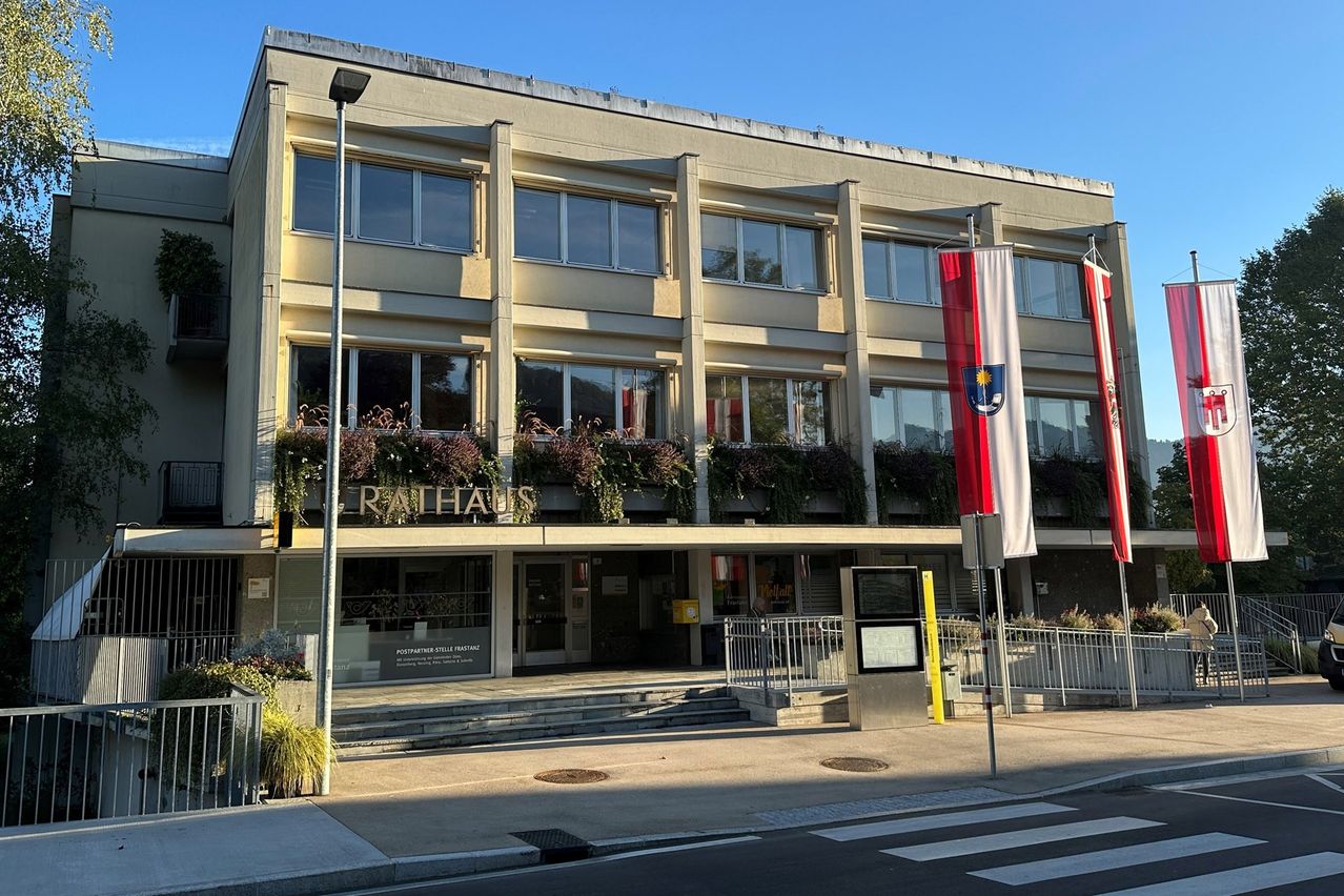 Ein Gemeindegebäude mit Glasfenstern und einem Schild mit der Aufschrift Rathaus. Davor weht eine rot-weiße Fahne auf einem Pfosten.
