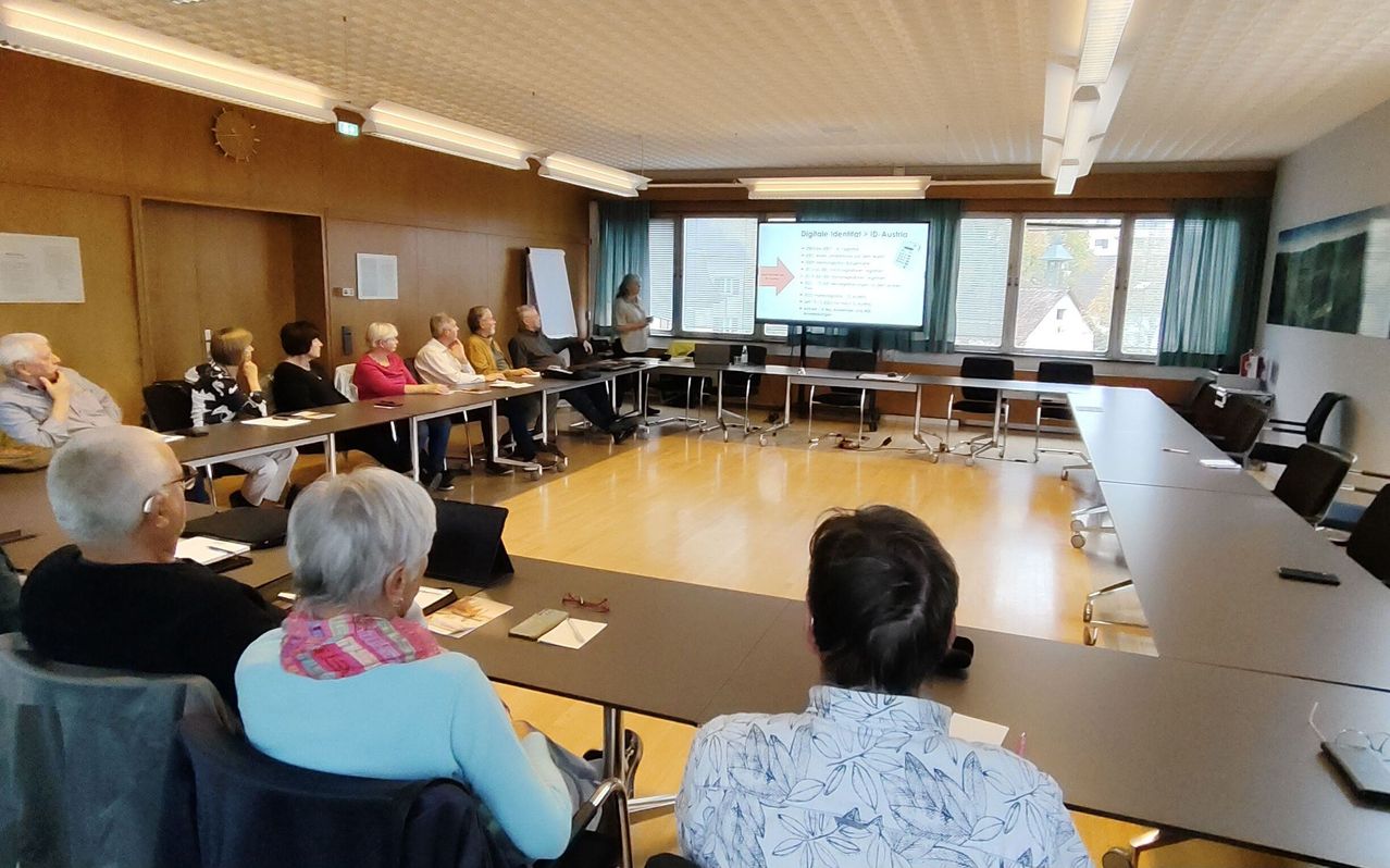 A group of elderly people are sitting around a table in a room. A woman is standing in front of a projector screen. She is giving a presentation. The room has wooden floors and is well lit.