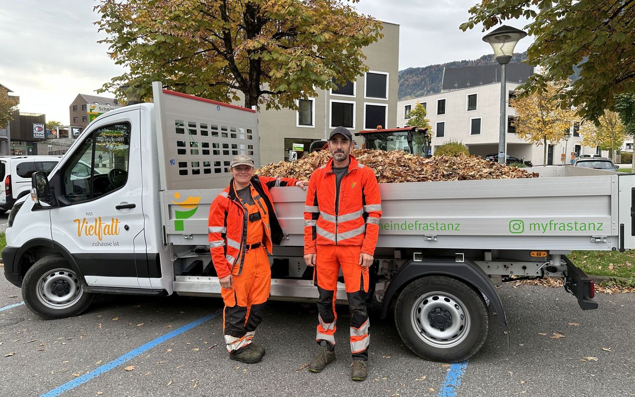 Two people in orange uniforms stand next to a truck filled with leaves, with a building and a tree in the background.