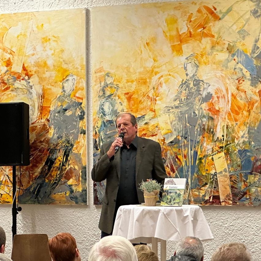 Ein älterer Mann spricht in ein Mikrofon an einem Podium mit zwei großen Gemälden hinter ihm.