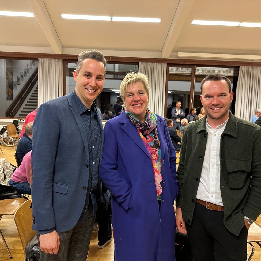 Drei Menschen stehen nebeneinander und lächeln. Sie sind bei einer Veranstaltung, wahrscheinlich einer Konferenz. Hinter ihnen stehen Stühle, Tische und sitzende und stehende Personen. Der Raum hat Glasfenster mit Vorhängen. Es gibt eine Treppe mit Bilderrahmen und eine Decke mit Lichtern.