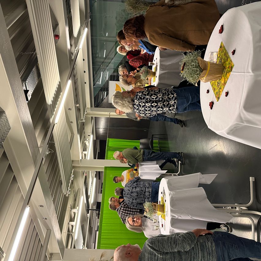 Eine Gruppe von Menschen versammelt sich in einem Raum, möglicherweise bei einer sozialen Veranstaltung. Der Raum hat eine grüne Kulisse und mit weißen Tüchern bedeckte Tische. Einige Menschen stehen und reden, während andere sitzen und essen. Der Raum hat eine weiße Decke mit Lichtern.