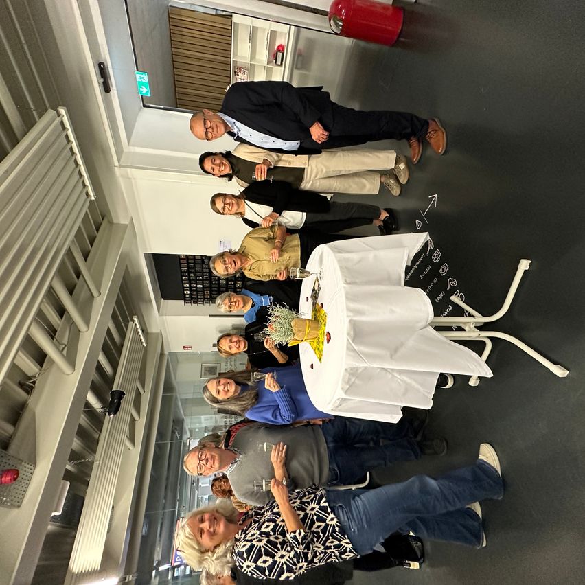 Eine Gruppe von Menschen steht lächelnd um einen Tisch und feiert möglicherweise ein Ereignis. Ein Mann hält ein Glas. Auf dem Boden befindet sich eine Zeichnung.