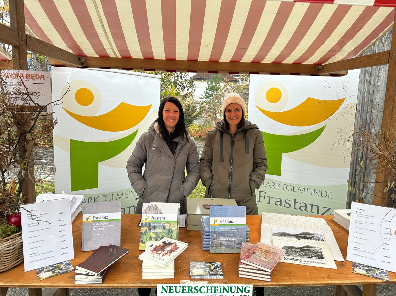 Zwei Frauen stehen hinter einem Tisch auf einem Markt. Sie tragen Wintermäntel und Mützen. Der Tisch ist mit Broschüren und Büchern bedeckt, und hinter ihnen sind Banner mit dem Text Frastanz.