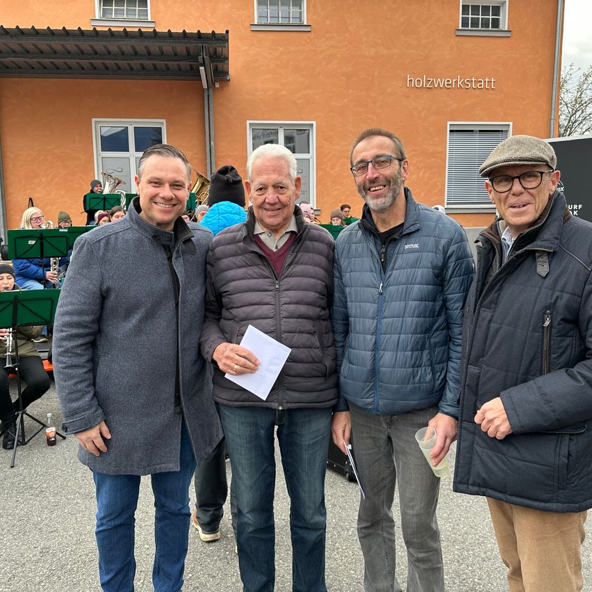 Vier Männer stehen lächelnd vor einem Gebäude. Der Mann in der Mitte hält ein Stück Papier in der Hand. Ein Musikorchester ist im Hintergrund zu sehen.