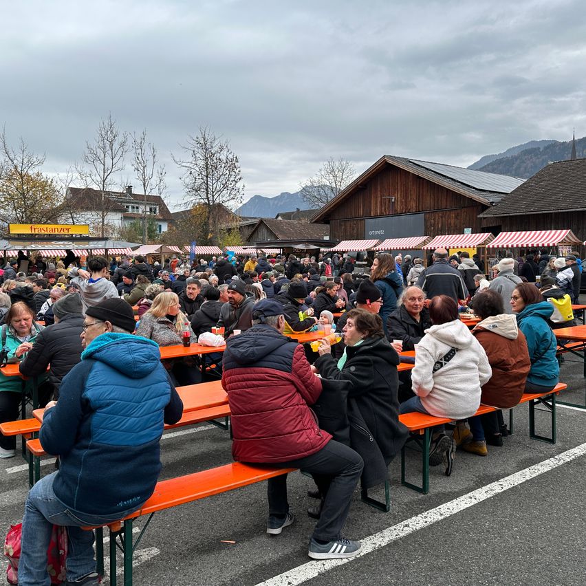 Eine Menschenmenge sitzt auf Bänken in einem Freiluftbereich, wahrscheinlich einem Markt, mit Bergen im Hintergrund. Einige tragen Mützen und Jacken, und es gibt Stände mit Schildern.