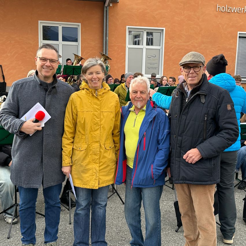 Vier Menschen stehen lächelnd vor einem Gebäude. Ein Mann hält ein Mikrofon. Dahinter versammelt sich eine Menge mit Musikinstrumenten. Das Gebäude trägt den Namen 'holzwerk' auf seiner Wand.