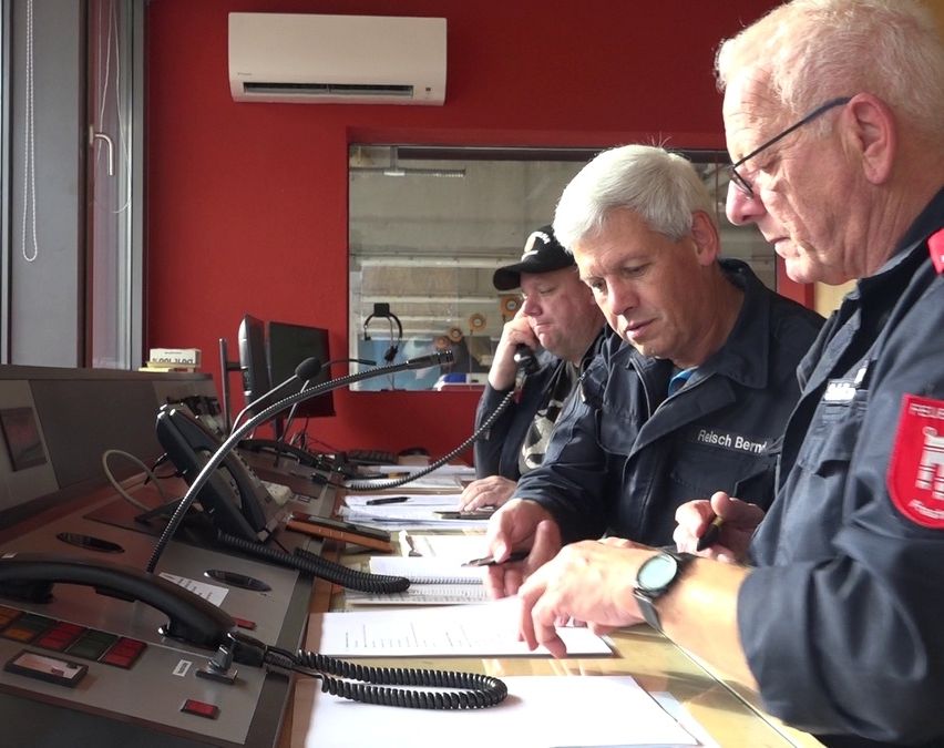 Drei ältere Männer arbeiten in einem Büro. Sie schauen sich Papiere auf einem Schreibtisch an. Einer schreibt. Der Raum hat eine rote Wand, Monitore und eine Klimaanlage.