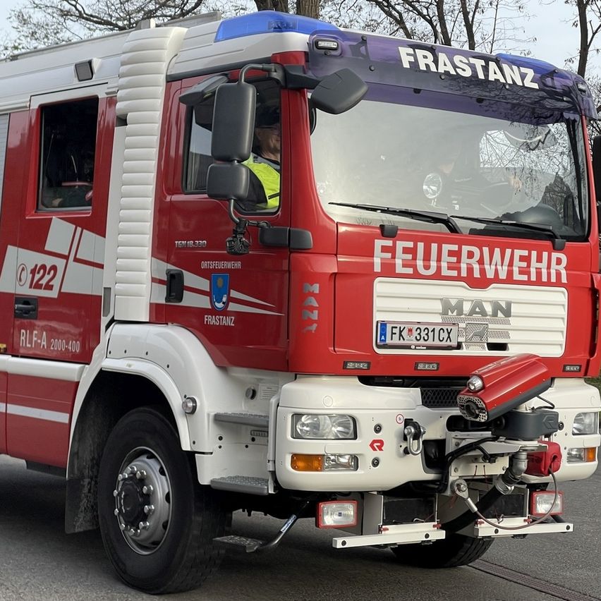 Ein roter Feuerwehrwagen mit 'FRASTANZ' und 'FEUERWEHR' auf der Front. Das Kennzeichen lautet 'FK 331CX'. Eine Person in einer gelben Weste sitzt auf dem Fahrersitz.
