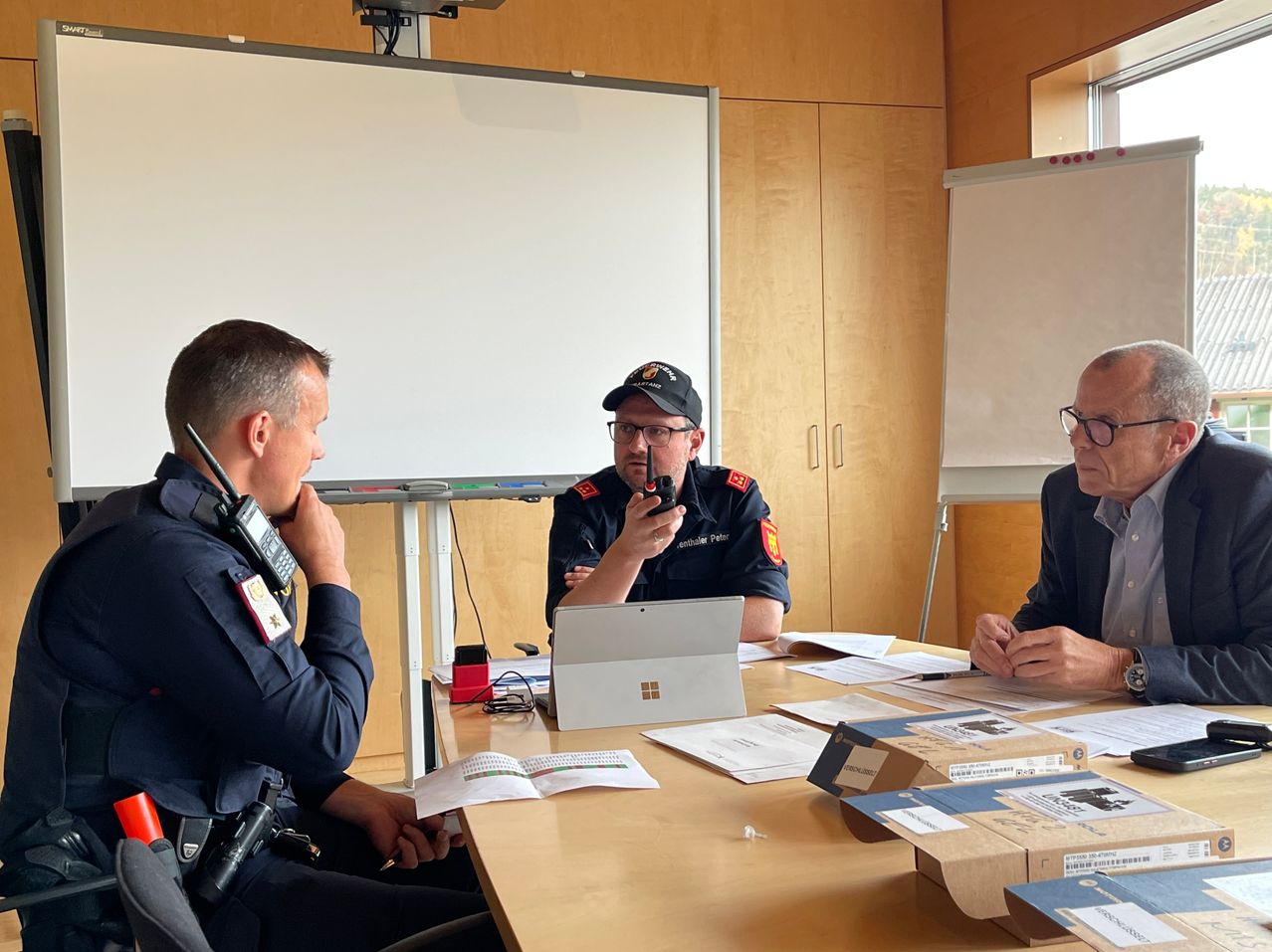 Drei Männer in Uniformen sitzen an einem Tisch, einer benutzt ein Walkie-Talkie, ein anderer einen Laptop und der dritte Papiere. Im Hintergrund sind eine Tafel und Schränke zu sehen.