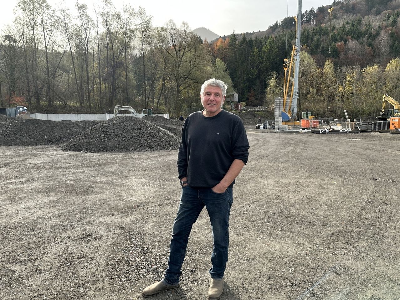 Ein Mann steht auf einem Schotterplatz mit einem Haufen Steine hinter ihm. Er trägt ein schwarzes Langarmhemd, Jeans und braune Schuhe. Er lächelt und schaut in die Kamera. Hinter ihm sind Baufahrzeuge und ein Kran. In der Ferne gibt es Bäume und Berge.