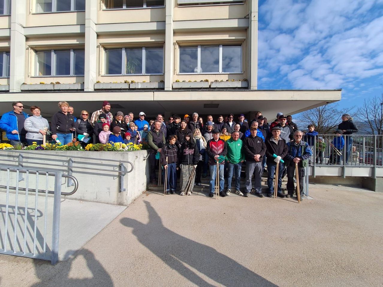 Eine Gruppe von Menschen in Winterkleidung und mit Stöcken posiert für ein Foto vor einem Gebäude mit Pflanzen und Geländern an einem bewölkten Tag.