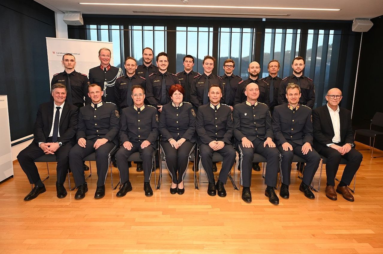 Eine Gruppe uniformierter Personen, wahrscheinlich Polizeibeamte, sitzen gemeinsam auf Stühlen in einem Raum mit großen Fenstern.