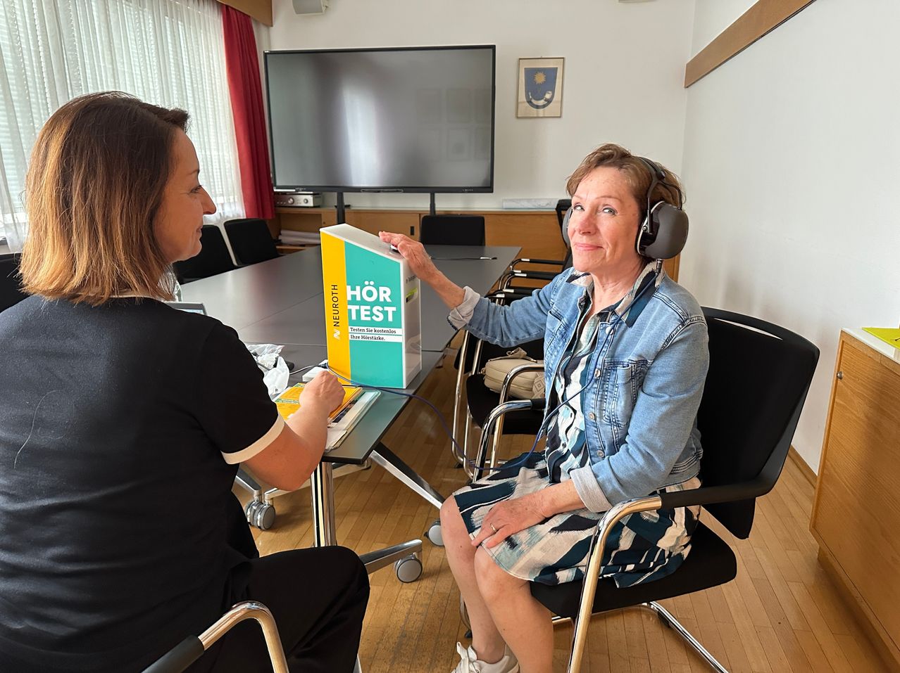 Two women in a room with a long table, one wearing headphones. A box labeled 'Hör Test' is on the table. Behind them, a monitor is mounted on the wall.