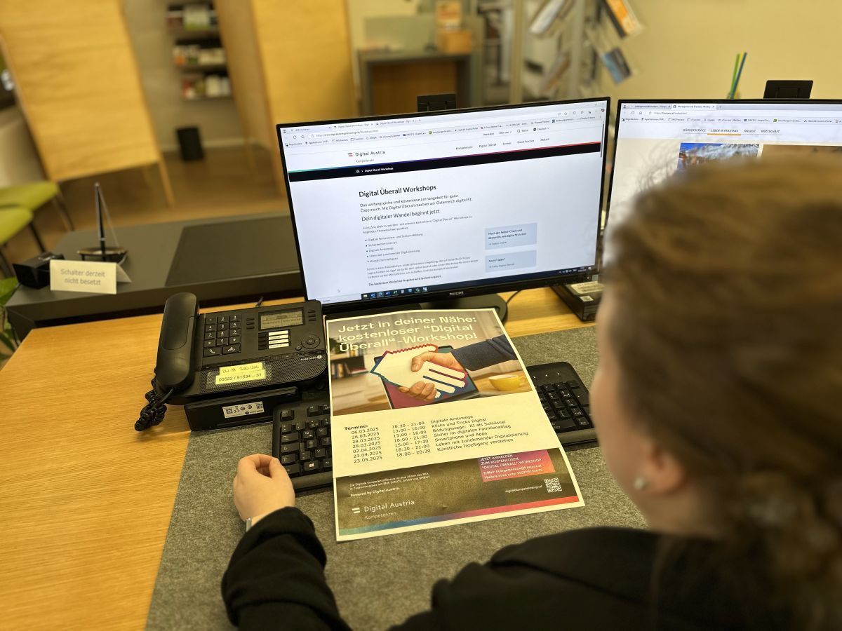 Eine Person sitzt an einem Schreibtisch mit Computer und Broschüre. Der Computerbildschirm zeigt eine Webseite über digitale Workshops an.