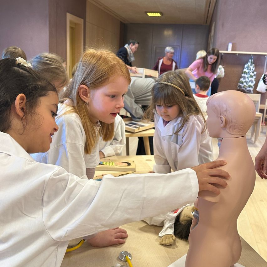 Eine Gruppe junger Mädchen in weißen Laborkittel übt Erste Hilfe an einem Mannequin. Andere Kinder und Erwachsene beobachten und lernen im Hintergrund.