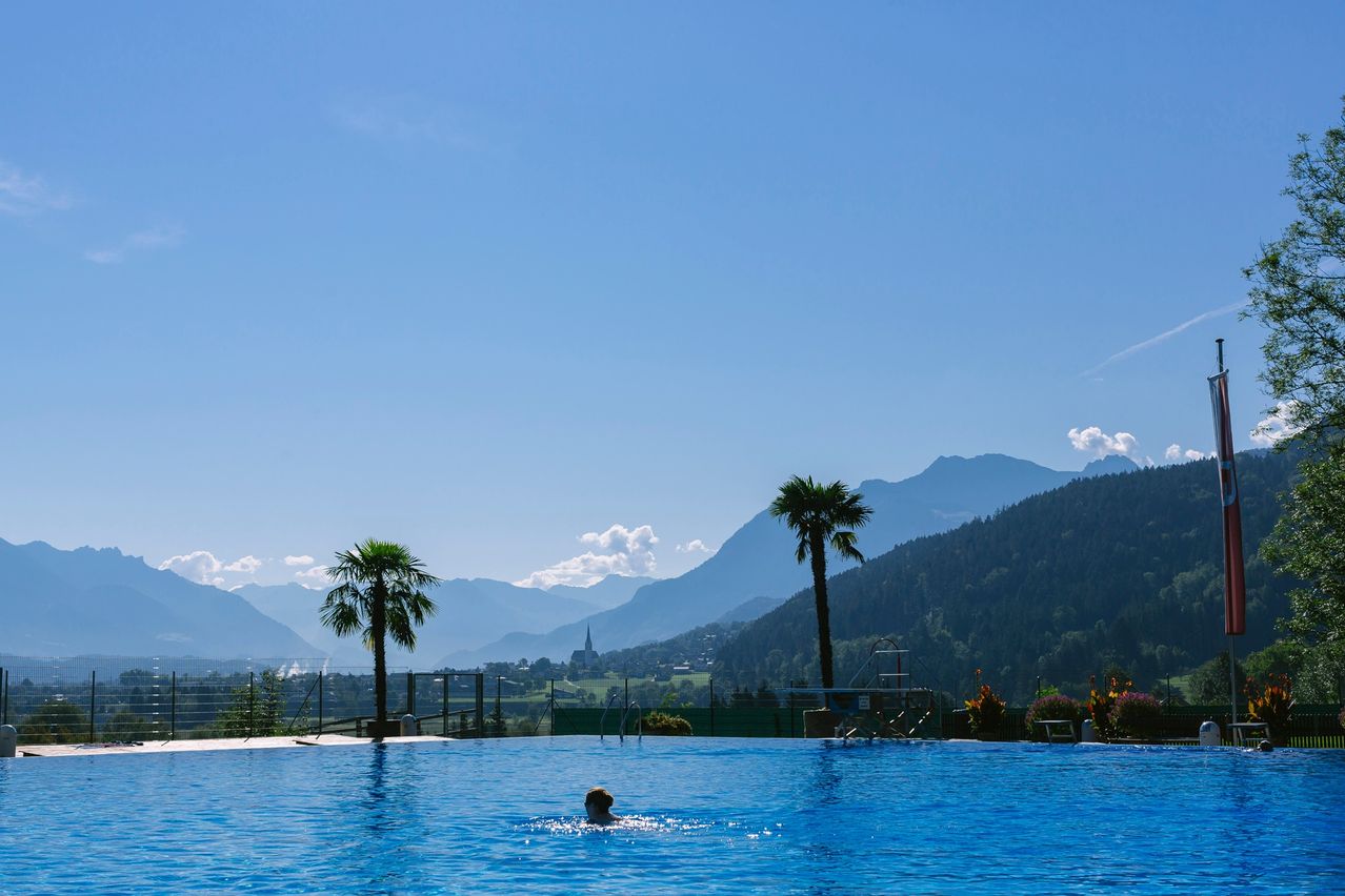Eine Person schwimmt in einem großen Freibad, umgeben von Palmen und Bergen. Der Himmel ist blau mit einigen Wolken.