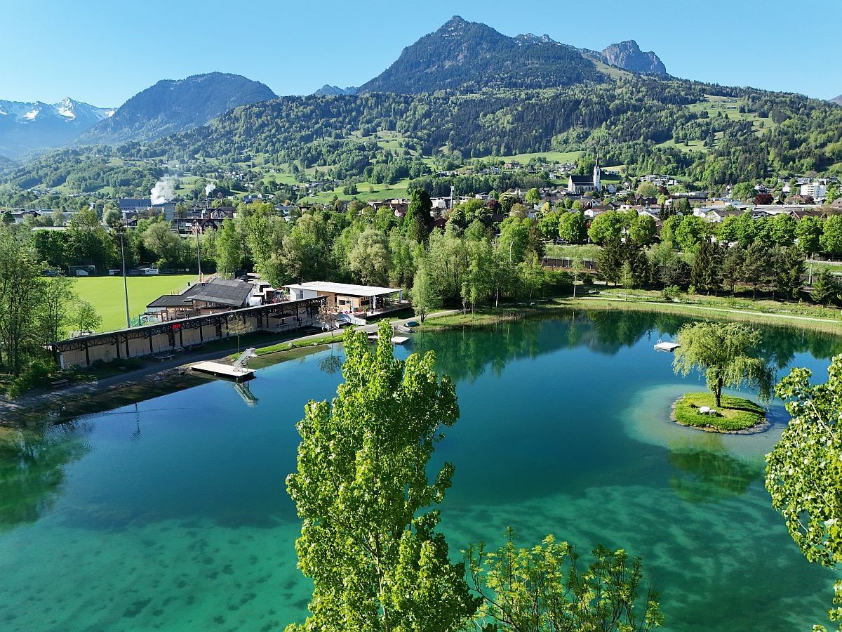 Ein ruhiger See, umgeben von üppigem Grün und Bergen. Ein Gebäude mit Dachterrasse befindet sich links. Bäume, ein Steg und eine kleine Insel im See.